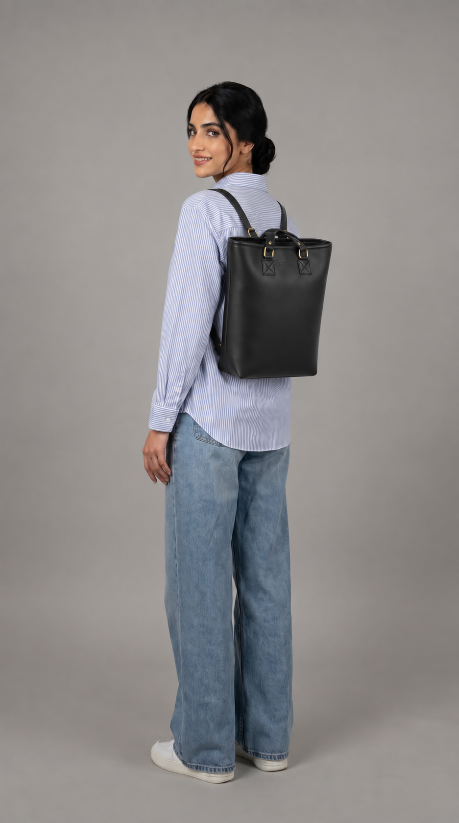 NOVYA Transit convertible women's tote bag and backpack in black
A woman wearing a light blue button-up shirt, wide-leg blue jeans, and white sneakers is shown from the back while wearing a black leather bucket backpack against a solid gray studio background.