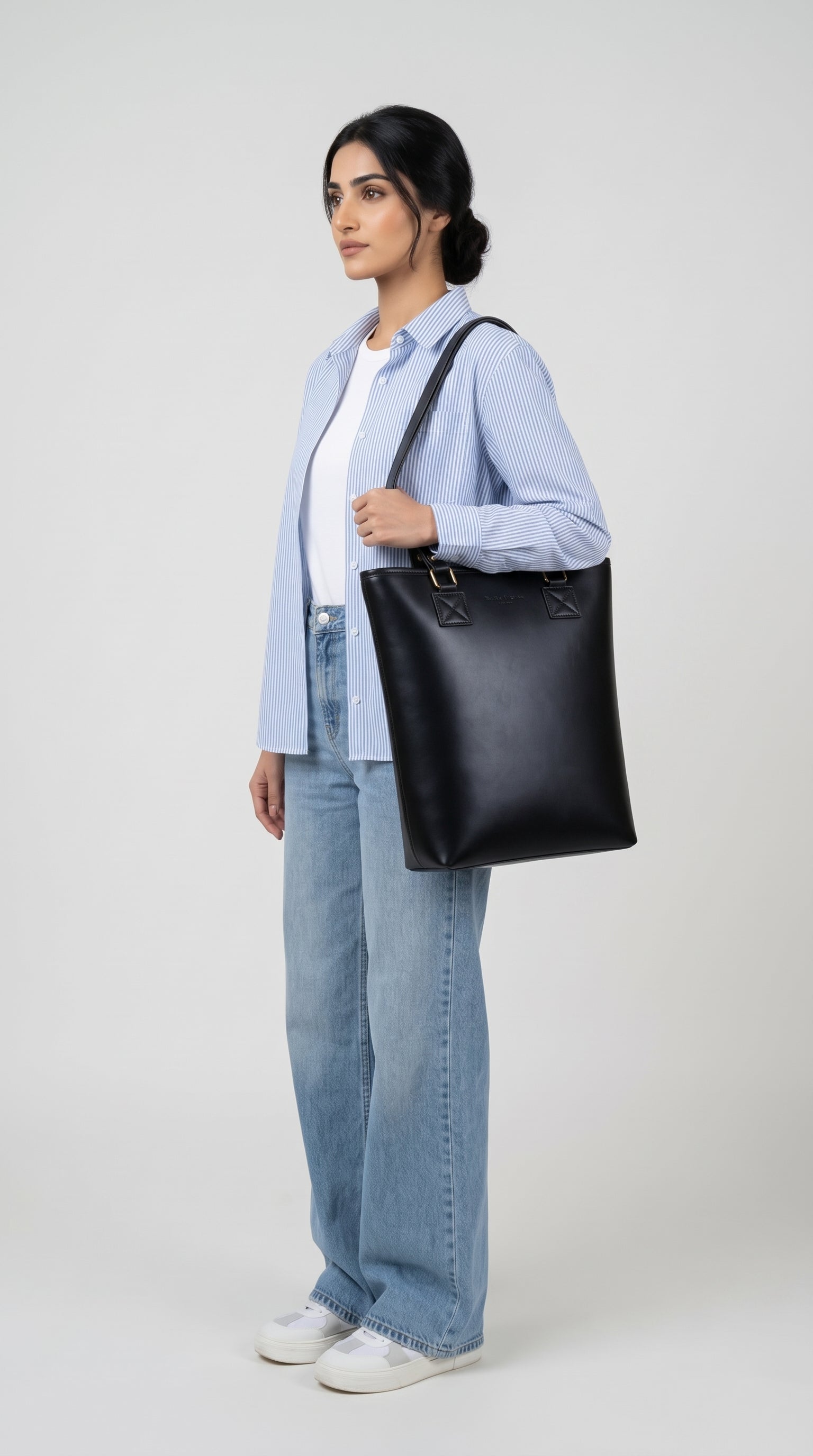 NOVYA Transit convertible women's tote bag and backpack in black.
A woman stands in profile wearing a light blue button-up shirt, white t-shirt, wide-leg light blue jeans, and white sneakers, while carrying a large black leather tote bag over her shoulder against a light gray studio background.